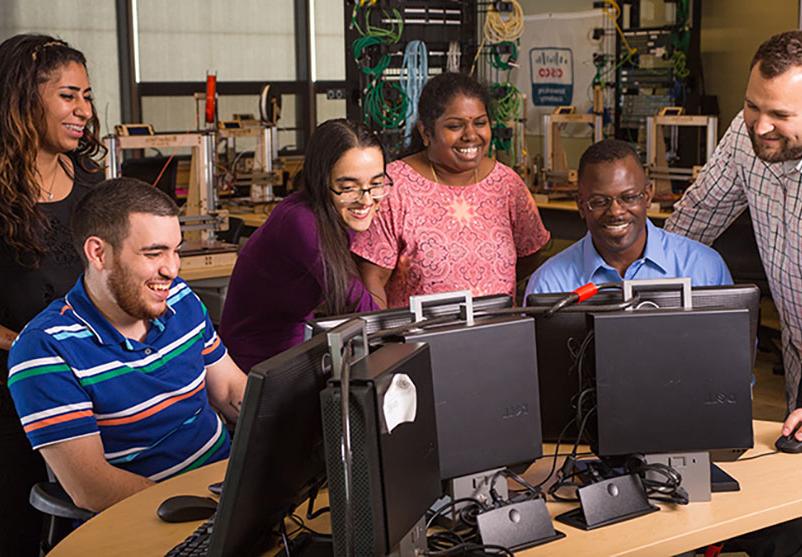 A group of students sitting and standing around two computers smiling.